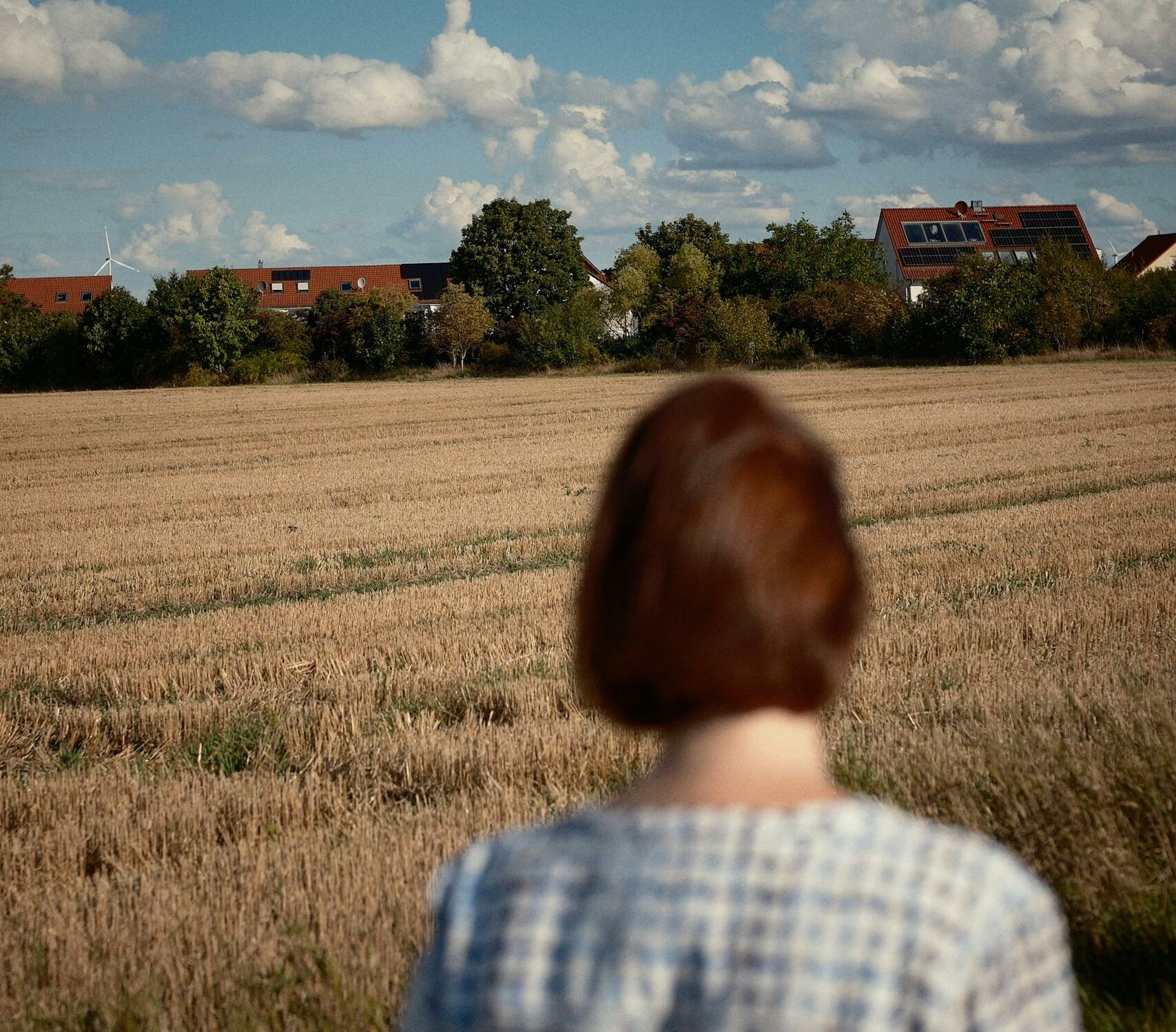 A woman stands in a field gazing towards a scenic rural landscape under a cloudy sky.